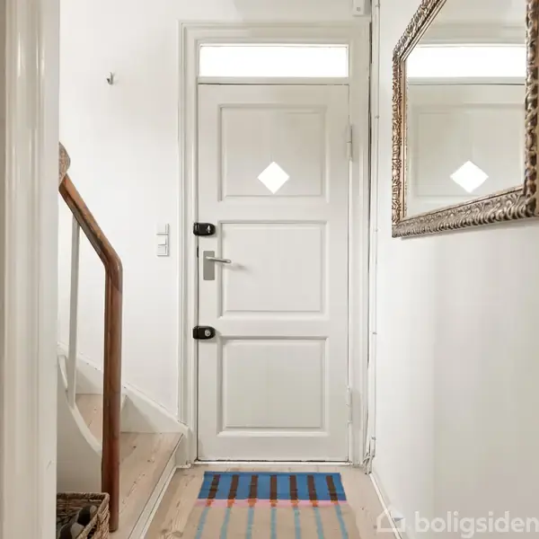 Dør åbner indad i en entré med hvidmalede vægge, trægulv, stribet tæppe og spejl på væggen. En trappe med trægelænder fører opad til venstre.
