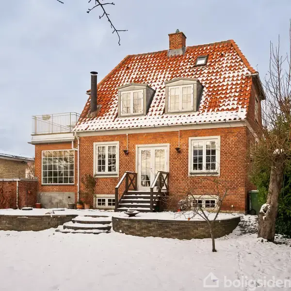 Et rødt murstenshus med rødt tegltag, har tre døre og vinduer på første sal. Omgivet af en snedækket have og en stentrappe, ligger det i et boligkvarter.