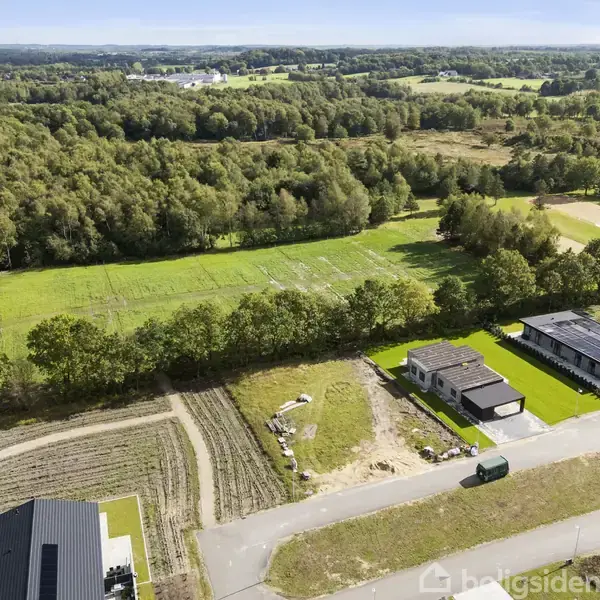 Et boligområde med et par enfamiliehuse under opførelse. Husene ligger langs en vej, omkranset af en stor, grøn skov og marker i baggrunden.