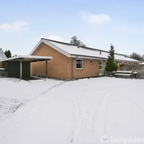 En enetages murstenshus står i et sneklædt landskab. Til venstre er en carport og til højre et hegn. Sne dækker jorden og omgiver huset.