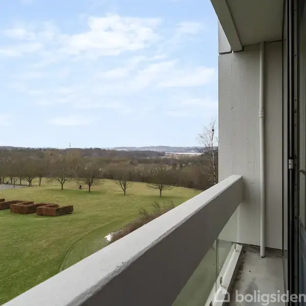 En balkon med glasrækværk giver udsigt over en grøn græsplæne med buske og træer i det fjerne under en blå himmel.