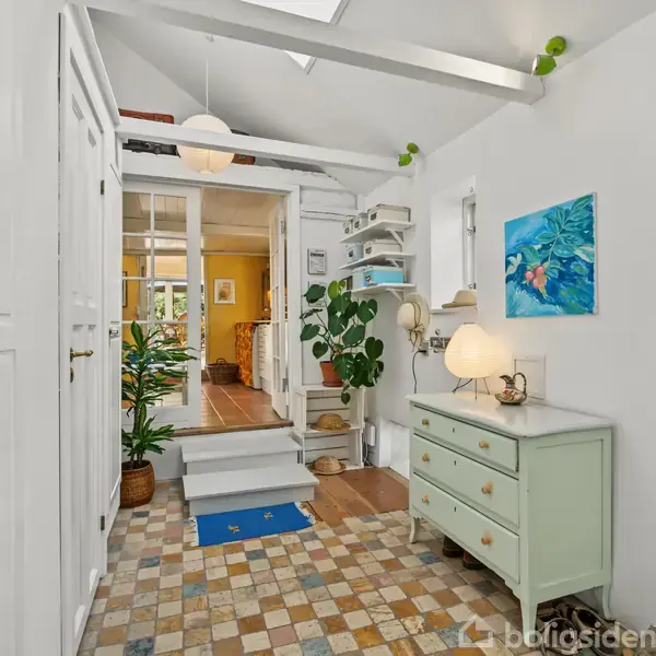 Entré med farverige klinker, hvor der står en mintgrøn kommode med lampe og dekorative genstande. Til venstre er der skabe, og i baggrunden ses en åben dør til et lyst rum.