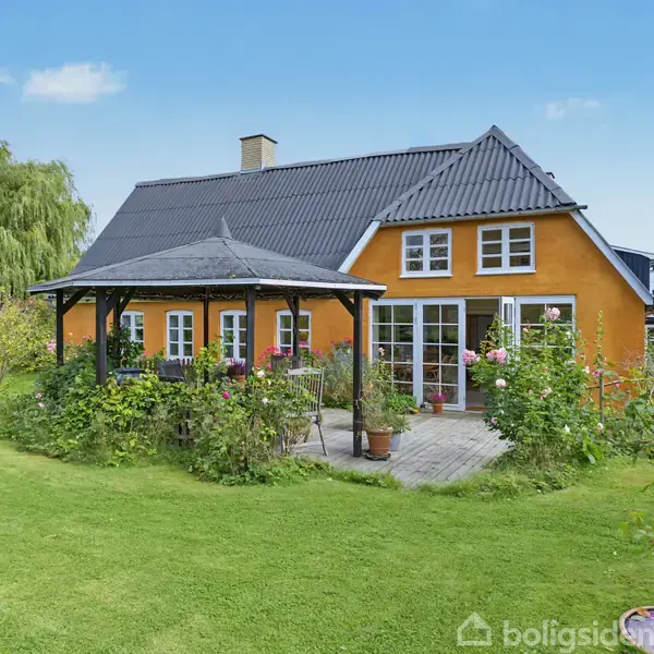 Et gult hus med sort tag står i en frodig have. Huset har en terrasse omgivet af blomster og træer, samt en åben pavillon ved siden af.