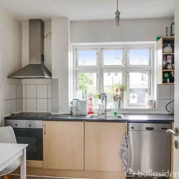 Køkken med komfur og emhætte ved siden af vask under et vindue, omgivet af hvide fliser og skabe. En opvaskemaskine står ved siden af, og et bord med stol ses til venstre. Ingen synlig tekst.