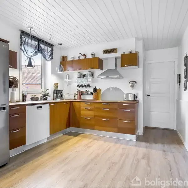 Køkken med brune og hvide skabselementer i et rummeligt rum. Køleskab og ovn i stål. Vindue med rullegardin. Trægulv og hvidt loft. Moderne indretning.