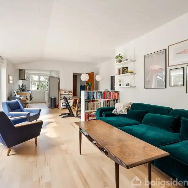 En stue med grøn sofa og træbord står i en lys, moderne lejlighed. Store vinduer slipper dagslys ind. Flere plakater hænger på væggen, og hylder rummer bøger og planter.