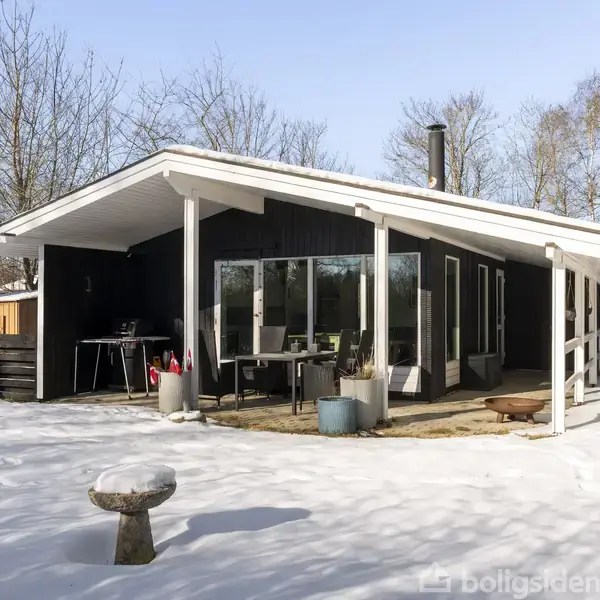 Et sort sommerhus med hvidt tag står stille i en snedækket have. Bord og planter er på terrassen, omkranset af træer og hegn.