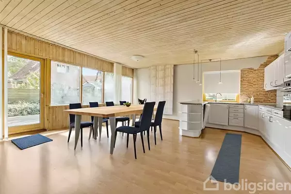 Spisebord med seks sorte stole står på trægulv i et lyst køkken med store vinduer. Køkkenet har hvide skabe, en vask, og naturstensvæg. Udendørsområde er synligt gennem vinduerne.
