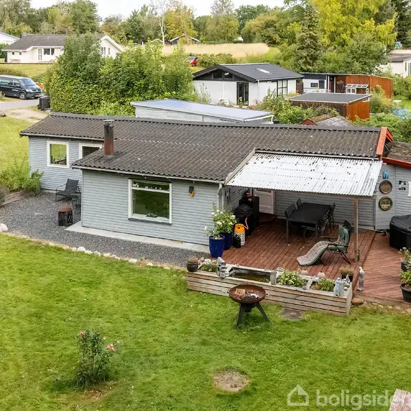 Et gråt hus med tagterrasse står i en grøn have, omgivet af forskellige planter og blomsterkrukker. Der er en åben gårdhave med udendørs møbler og en grill.