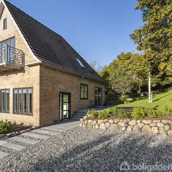 Et murstenshus med gavltag står på en grusbelagt grund. Huset har store vinduer og balkon. Omkringliggende træer og en græsplæne bidrager til et frodigt miljø.