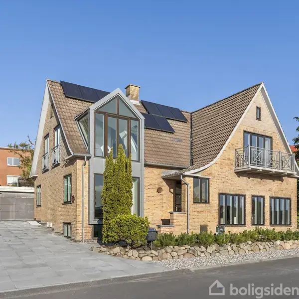 Et moderne hus med solpaneler på taget står i et boligområde. Bygningen har store vinduer og en carport. Omgivet af en flisebelagt have og nabobygninger.