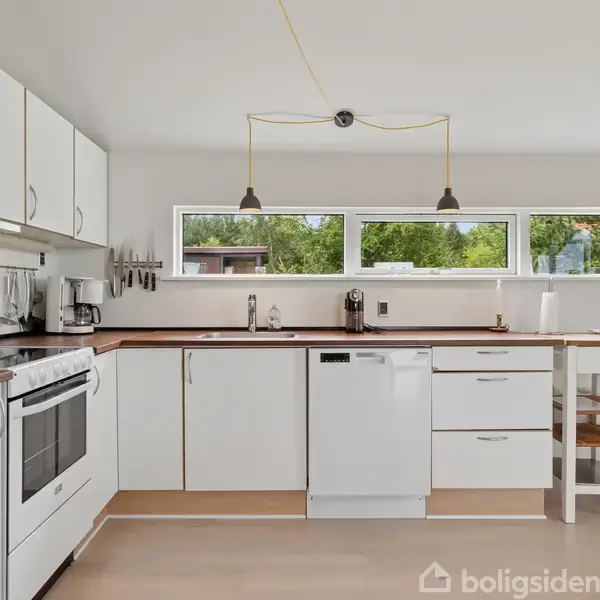 Køkken med hvide skabe og apparater, såsom komfur og opvaskemaskine, placeret langs væggen. To hængelamper ovenover. Vinduer giver udsigt til grønt landskab udenfor.