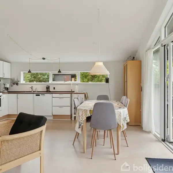 Et lyst køkken-alrum rummer et spisebord med stole, omgivet af et moderne køkken med hvide skabe. En stor glasskydedør åbner ud til en terrasse med udsigt til grøn natur.