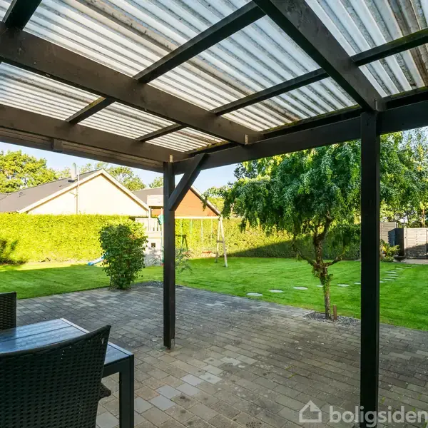 En overdækket terrasse med træbord og stole står på flisebelagt område. I baggrunden ses en grøn have med træer og hække, omgivet af et roligt boligområde.