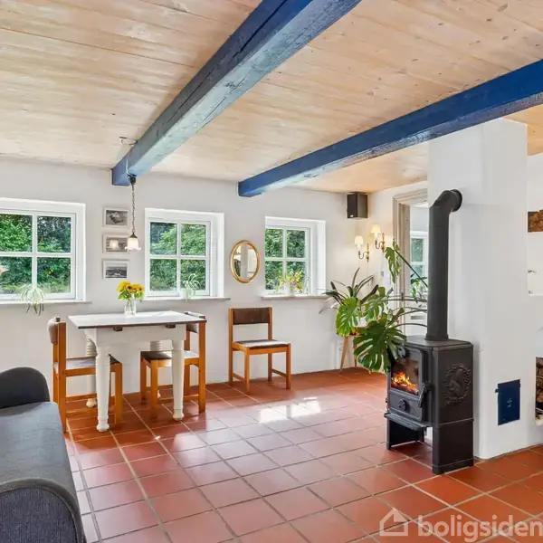 Stue med pejseovn i hjørnet, omgivet af møbler som sofa og spisestuesæt. Rummets hvide vægge og træloft med blå bjælker skaber en lys atmosfære med planter og vinduer med udsigt til natur.