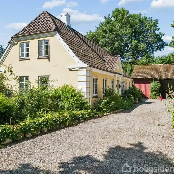 Et gult hus med brunligt tag står omgrænset af frodig vegetation. En grusvej fører op til huset. I baggrunden ses et tilhørende skur under blå himmel.