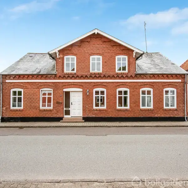 En rødstensbygning med hvide vinduer og en hvid dør står stille på en stille gade. Bygningen har gråt tegltag og omgives af to andre huse med rødt og hvidt tag.
