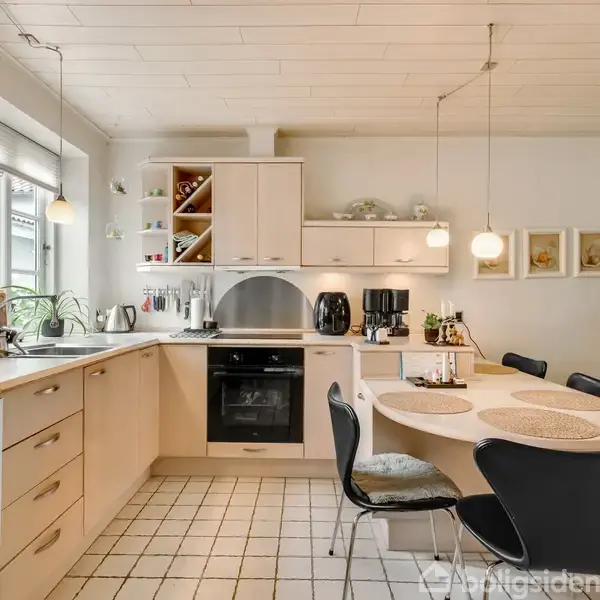Køkken med hvide skuffer og skabe, kogeplade og ovn centralt. Rundt bord med stole til højre. Moderne lamper hænger fra loftet. Vindue til venstre slipper lys ind.