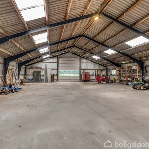 En stor, tom lagerhal rummer forskellige landbrugsmaskiner og værktøj, opstillet langs væggene. Loftet er højt og har ovenlys. Rummet bærer præg af at være arbejdsrelateret.