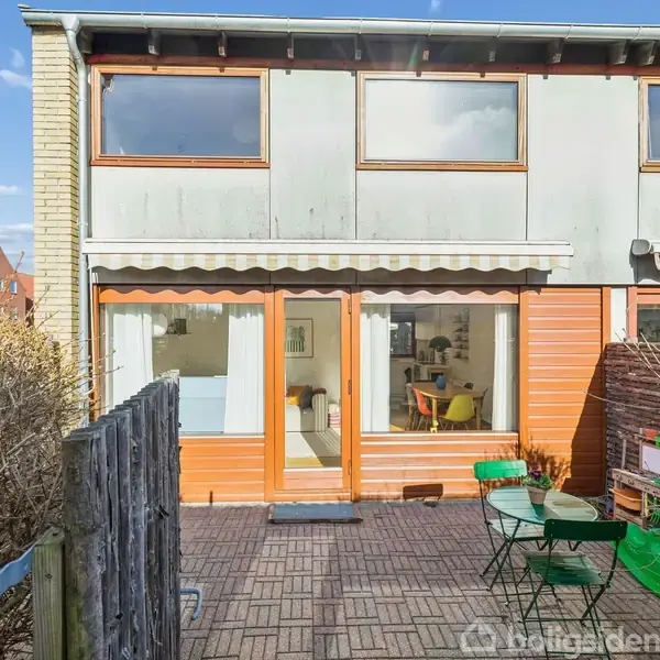 Rækkehus med hvid og orange facade, to etager, store vinduer og en terrasse med havemøbler. Omkranset af hæk og plankeværk, beliggende i et beboelseskvarter med klar himmel.