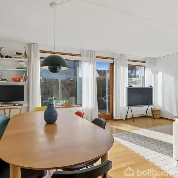 Spisebord med vase står i stue med store vinduer og lyse gardiner. Til venstre ses hylde med diverse genstande og en skærm. I hjørnet står et TV på stativ.