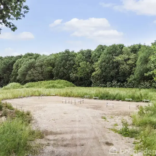 En sandet byggeplads med markerede fundamentrør står urørt. Omkring den er frodig vegetation og tætte træer, der danner en grøn baggrund under en klar blå himmel.