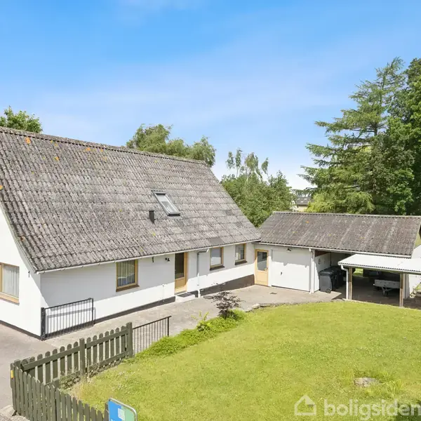 Hvidt hus med gråt sadeltag har flere vinduer og en tilknyttet carport. Det står i en have med grøn græsplæne, omkranset af hegn og træer i baggrunden.