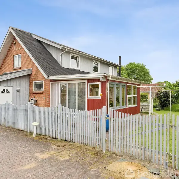 Et rødt murstenshus med to etager står stille omgivet af en grøn have og hvidt stakit. Foran huset er der en flisebelagt indkørsel under en klar blå himmel.