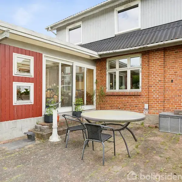 En rund havebord med to stole står på en terrasse ved et hus med røde mursten og en tilbygning i træ. Vinduerne giver udsigt til haven.