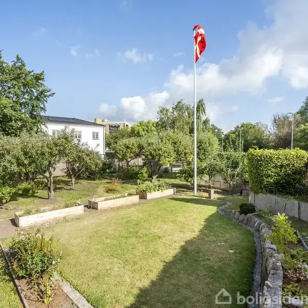 Flagstang med dannebrog står i solrig have omgivet af grønne buske og træer. En hvid bygning kan ses i baggrunden under en blå himmel med spredte skyer.