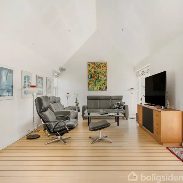En stue med et sofabord står i midten omgivet af to grå lænestole og en sofa. Et fladskærms-TV står på et træmøbel til højre. Væggene har kunst og store glasdøre udgør indgangen.