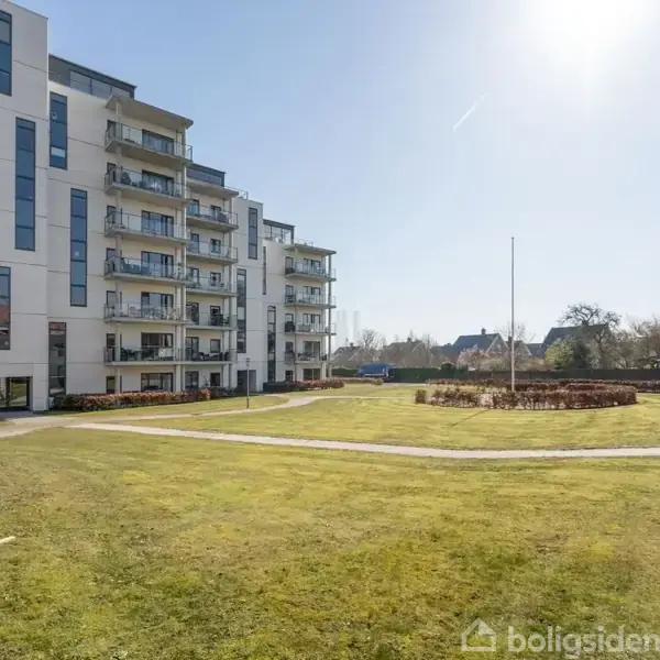 Flereetages bygning med altaner står til venstre. Græsplæne og stier udgør en gårdhave. Træer og buske ses i baggrunden under en klar, blå himmel.