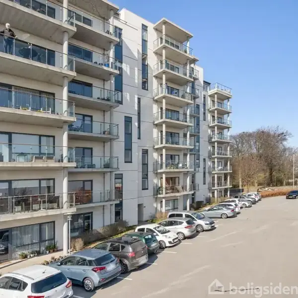 En moderne, fleretagers beboelsesejendom står ved siden af en parkeringsplads fyldt med biler. En person står på en altan overfor træer i baggrunden.