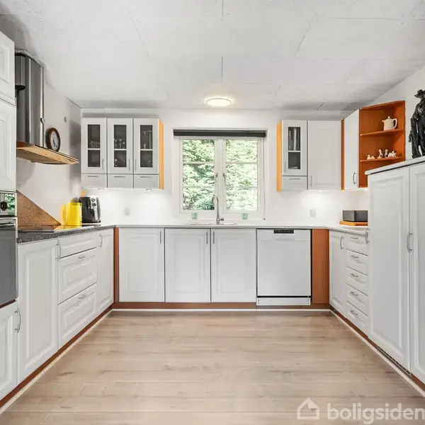 Et moderne køkken med hvide skabe og stålapparater, herunder ovn og opvaskemaskine. En stor vindue bagvæggen giver udsigt til træer udenfor. Trægulve og dekorative figurer på hylderne.