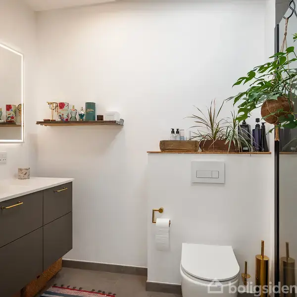Badeværelse med mørk vaskeskab og spejl på venstre væg, vægmonteret toilet i midten. Hylder med dekorative genstande og planter på væggen. Hængende planter ved bruseniche på højre side.
