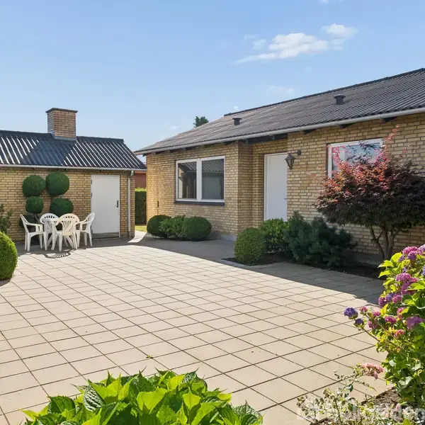 Bungalow i gule mursten står ved en flisebelagt terrasse. En plastikhavemøbelgruppe står til højre. Haven har grønne buske og blomster. Solens lys falder på den åbne gårdsplads.