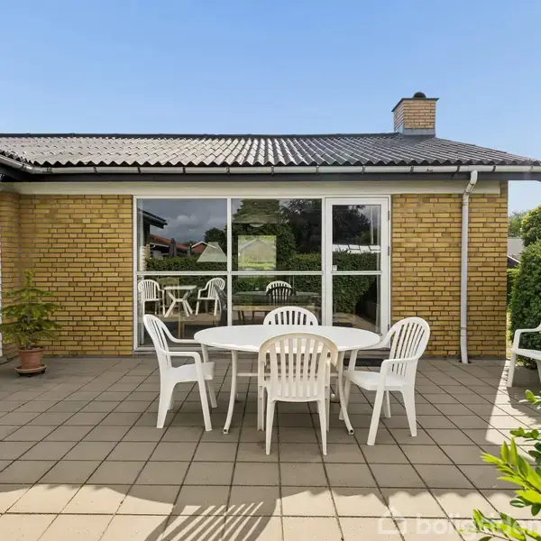En hvid plastikhavemøbelgruppe med fire stole står på en flisebelagt terrasse foran et gult murstenshus med glasparti og skorsten. Grønne buske omgiver området.