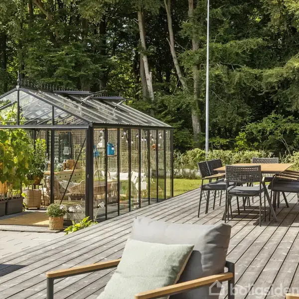 Et drivhus fyldt med planter står på en træterrasse ved siden af et spisebord med stole. Omgivet af frodig skov skaber det en idyllisk haveoplevelse.