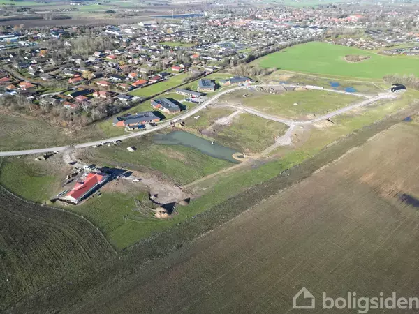 Luftfoto viser en nyudviklet boligområde med huse i forskellige konstruktionstrin omkring en lille sø; omgivelserne består af landbrugsjord og en nærliggende by i baggrunden.