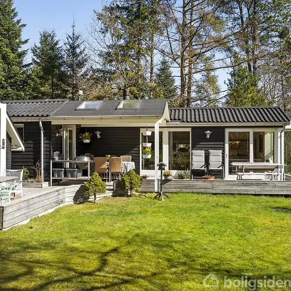 Et sort, træbeklædt sommerhus med flere terrasser placerer sig i en grøn have omgivet af høje træer. Møbler og planters pynter terrasserne, mens solen skinner klart.