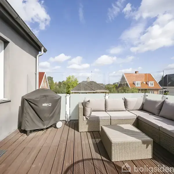 En udendørs terrasse med en L-formet sofa og et sofabord på trægulv. En grill står dækket ved væggen. Omkring er der udsigt over hustage under en blå himmel.