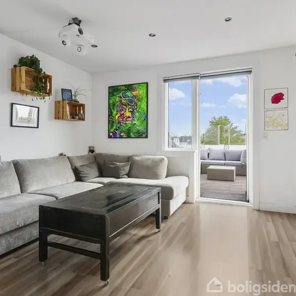 Grå sofa står mod en hvid væg med farverige malerier og træhylder. Sofaen vender mod et sort bord. En dør fører ud til en terrasse med siddepladser.