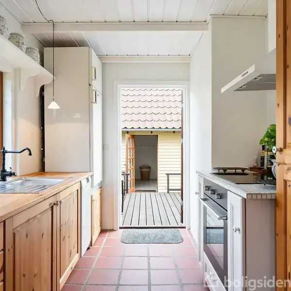 Et køkken med træskabe og komfur, hvor en dør fører ud til en terrasse. Store vinduer til venstre slipper lys ind, og der ses grønt udenfor.
