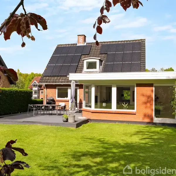Hus med solpaneler står stille i en solrig have med græsplæne, flankeret af en terrasse med havemøbler. Omkringliggende grønne træer rammer scenen ind.