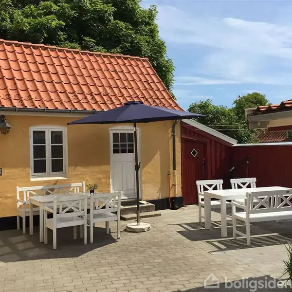 Gul husbygning med rød tegltag; to hvide døre. To hvide sæt havemøbler står i gårdspladsen; parasol skygger, omgivet af rødt plankeværk og grønne trætoppe.