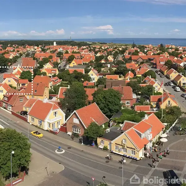 Røde tage dækker ensartede huse i en by med brostensbelagte gader og frodige træer. Bygningerne omkranser en central vej, mens havet kan skimtes i baggrunden under en klar blå himmel.