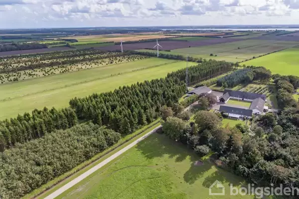 Gården ligger omgivet af marker og skove med to vindmøller i horisonten. En lang vej fører til bygningen, der er omringet af træer.
