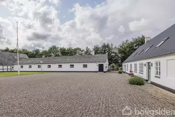 En gårdsplads med grusbelægning omgivet af to hvide landbygninger med sorte tage. Flagstang og lamper står i gården, omkranset af træer under en skyet himmel.