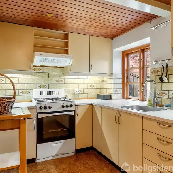 Komfur med emhætte placeret i hjørnekøkken, omgivet af lysebrune skabe og skuffer. Vinduet over vasken giver udsigt til en murstensbygning. På træbordet er en kurv.