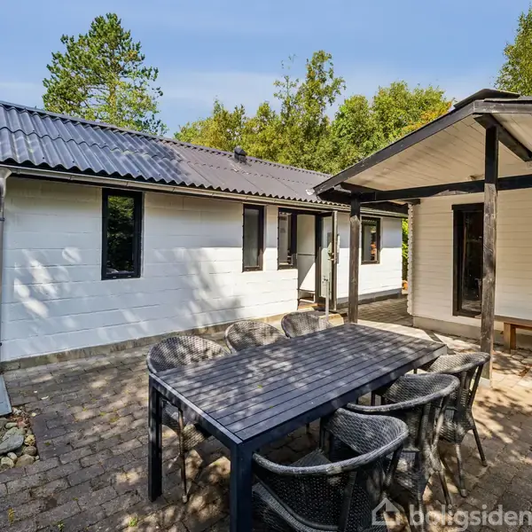 Huset har en hvid facade og sort tag, placeret i en have med frodige grønne træer. På terrassen står et bord med stole, omgivet af fliser.